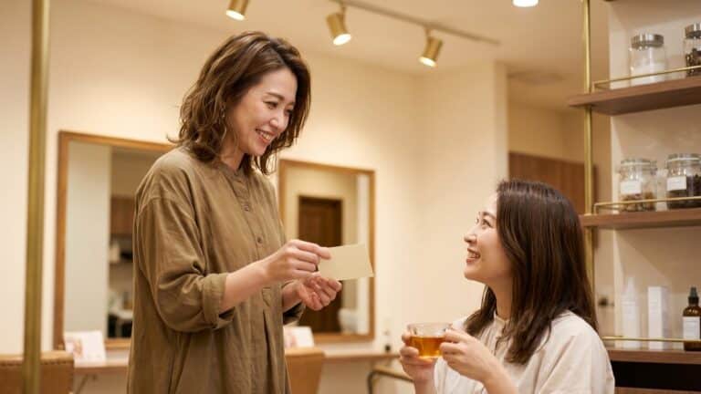Female hairstylist handing a referral card to a happy salon client at a styling station, warm salon lighting